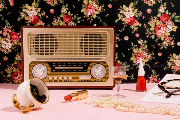 Retro tarzı radyo, bir kahve fincanı, kısa içecek bardağı, güzellik malzemeleri, bir kitap ve okuma gözlükleri, yaratıcı nostalji düzeni, arka planda çiçek duvar kağıdı..