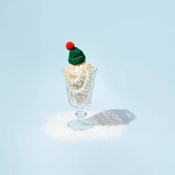 Creative winter holidays inspired dessert made of pearls in retro wine glass with warm deep green woolen hat on top. Icy blue background.