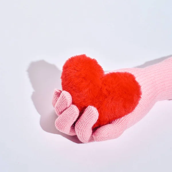 A woman's hand in a pink woolen glove holds a red fur heart, the idea of care, love, kindness.