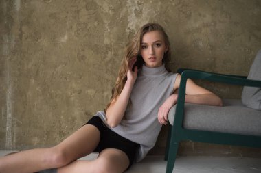 Siyah saçlı, bej bluzlu ve kot pantolonlu genç bir kız gri bir stüdyoda bir sandalyede oturuyor. 