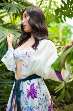 Beyaz kimonolu, mavi elbiseli yeşil ağaçların arkasında poz veren genç Asyalı kadın.