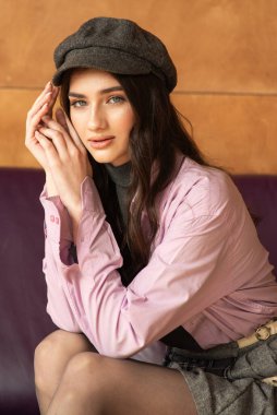 Pembe elbiseli, uzun saçlı ve kafasında şapka olan güzel esmer manken kahverengi ahşap arka planda kameraya bakıyor..