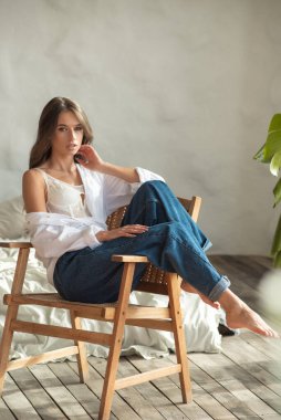 Beyaz gömlekli, kot pantolonlu, sandalyede oturan ve stüdyoda poz veren güzel bir kız..