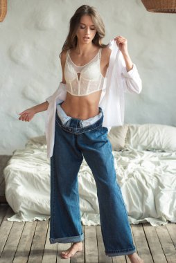 Beyaz iç çamaşırlı ve kot pantolonlu genç kadın evde poz veriyor.