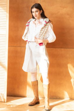 Stüdyoda beyaz gömlekli, kahverengi duvarlı, beyaz çizmeli genç bir kadın..