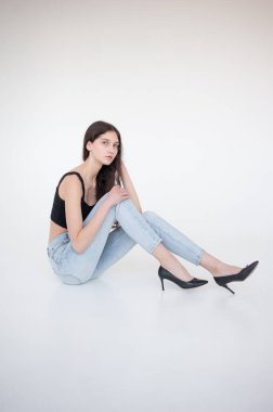 Güzel bir kadın, beyaz bir zeminde oturuyor. Kot pantolon ve siyah ayakkabılarla.