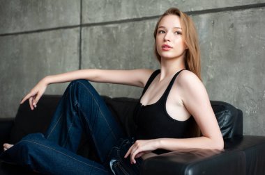 Uzun kahverengi saçlı, siyah tişörtlü ve kot pantolonlu güzel bir kız pencerenin yanında gri bir arka planda oturuyor..