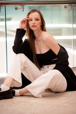 Beyaz gömlekli ve siyah pantolonlu genç kız yerde oturuyor.