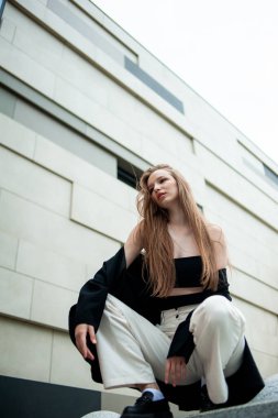 Beyaz elbiseli ve siyah ceketli kız merdivenlerde oturuyor ve kameraya bakıyor.