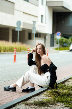 Binanın yanındaki bankta oturan beyaz gömlekli ve siyah kotlu güzel esmer kadın.