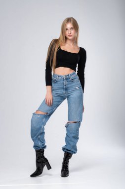 Sıradan kot pantolon ve deri pantolon giyen, eli kamerada duran güzel sarışın kızın tam boy portresi. Gri stüdyo arka planı.