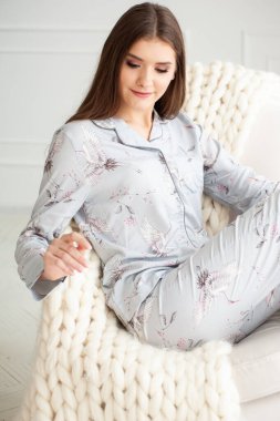 Pembe pijamalı, uzun saçlı ve mavi kazaklı güzel, genç esmer kadın beyaz yastıklı beyaz bir kanepede oturuyor..