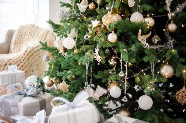 Süslü, altın ve beyaz bir ağaç, sarılı hediyeler, klasik bej bir kanepe ve rahat örgü battaniyeli bir koltuk. Parlak beyaz duvarlar şenlikli, minimalist bir ortam yaratır.