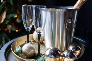 Stylish New Year celebration setup with a champagne bottle in a silver ice bucket, two elegant glasses, gold ornaments, and pine branches on a golden tray, set against a dark, luxurious background clipart