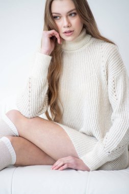 Büyük beden bir süveter ve diz boyu çoraplı genç bir kadın beyaz bir duvara karşı poz veriyor. Uzun, dalgalı saçları gülümseyen yüzünü çerçeveler, minimalist bir ortamda sıcaklığı ve rahatlığı yayar.