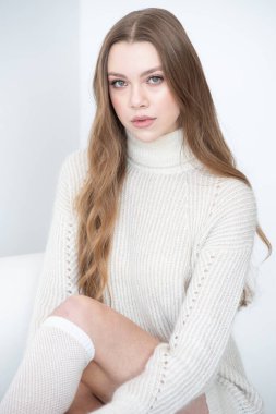 Büyük beden bir süveter ve diz boyu çoraplı genç bir kadın beyaz bir duvara karşı poz veriyor. Uzun, dalgalı saçları gülümseyen yüzünü çerçeveler, minimalist bir ortamda sıcaklığı ve rahatlığı yayar.