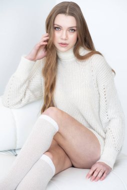 Büyük beden bir süveter ve diz boyu çoraplı genç bir kadın beyaz bir duvara karşı poz veriyor. Uzun, dalgalı saçları gülümseyen yüzünü çerçeveler, minimalist bir ortamda sıcaklığı ve rahatlığı yayar.