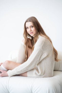 Büyük beden bir süveter ve diz boyu çoraplı genç bir kadın beyaz bir duvara karşı poz veriyor. Uzun, dalgalı saçları gülümseyen yüzünü çerçeveler, minimalist bir ortamda sıcaklığı ve rahatlığı yayar.