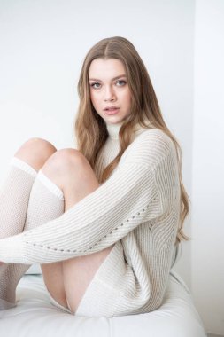 Büyük beden bir süveter ve diz boyu çoraplı genç bir kadın beyaz bir duvara karşı poz veriyor. Uzun, dalgalı saçları gülümseyen yüzünü çerçeveler, minimalist bir ortamda sıcaklığı ve rahatlığı yayar.