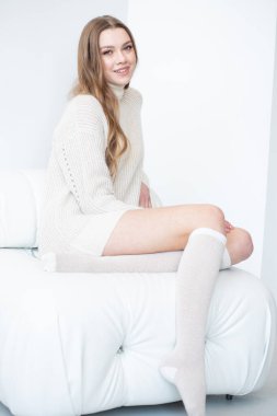 Büyük beden bir süveter ve diz boyu çoraplı genç bir kadın beyaz bir duvara karşı poz veriyor. Uzun, dalgalı saçları gülümseyen yüzünü çerçeveler, minimalist bir ortamda sıcaklığı ve rahatlığı yayar.