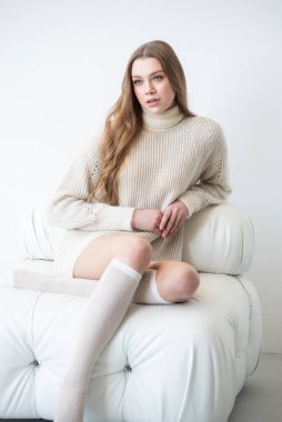 Büyük beden bir süveter ve diz boyu çoraplı genç bir kadın beyaz bir duvara karşı poz veriyor. Uzun, dalgalı saçları gülümseyen yüzünü çerçeveler, minimalist bir ortamda sıcaklığı ve rahatlığı yayar.