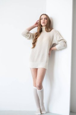 A young woman in a cozy beige oversized sweater and knee-high socks poses against a white wall. Her long, wavy hair frames her smiling face, exuding warmth and comfort in a minimalist setting clipart