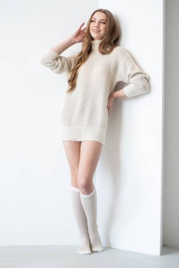Büyük beden bir süveter ve diz boyu çoraplı genç bir kadın beyaz bir duvara karşı poz veriyor. Uzun, dalgalı saçları gülümseyen yüzünü çerçeveler, minimalist bir ortamda sıcaklığı ve rahatlığı yayar.