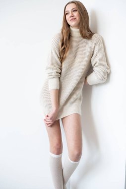 Büyük beden bir süveter ve diz boyu çoraplı genç bir kadın beyaz bir duvara karşı poz veriyor. Uzun, dalgalı saçları gülümseyen yüzünü çerçeveler, minimalist bir ortamda sıcaklığı ve rahatlığı yayar.