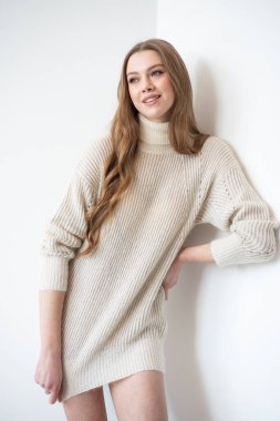 Büyük beden bir süveter ve diz boyu çoraplı genç bir kadın beyaz bir duvara karşı poz veriyor. Uzun, dalgalı saçları gülümseyen yüzünü çerçeveler, minimalist bir ortamda sıcaklığı ve rahatlığı yayar.