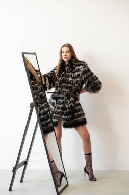Şık bir kadın siyah-beyaz sahte kürk manto ile poz veriyor koyu bir deseni var, siyah iç çamaşırı tarzı bir bluz giyiyor. Uzun, düz, açık kahverengi saçları ve kendine güveni, şık ve gergin görünmesini sağlıyor.