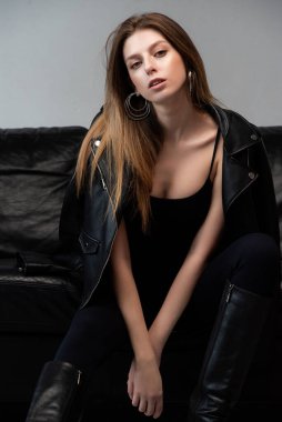 Deri ceketli ve siyah deri ayakkabılı genç kız siyah kanepenin yanında poz veriyor.