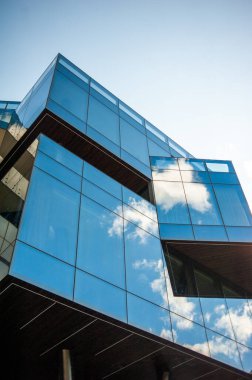 Cam paneller ve parlak tasarım içeren modern bir binanın mimari yakın çekimi. Aynalı yüzeyler gökyüzünü yansıtıyor, şehir planlamada yenilik ve zarafeti vurguluyor.