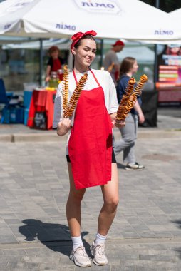 Önlüklü ve bantlı çekici genç bir kadın elinde patates şişi tutarken sokak yemeklerine gülümsüyor. Açık şehir fuarında ferahlatıcı limonata kavanozları ve şenlikli yaz atmosferi.