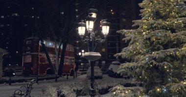 Noel akşamı şiddetli kar yağışı, bir Noel ağacının arka planında, parlak bir fener ve çift katlı kırmızı otobüs. 4K