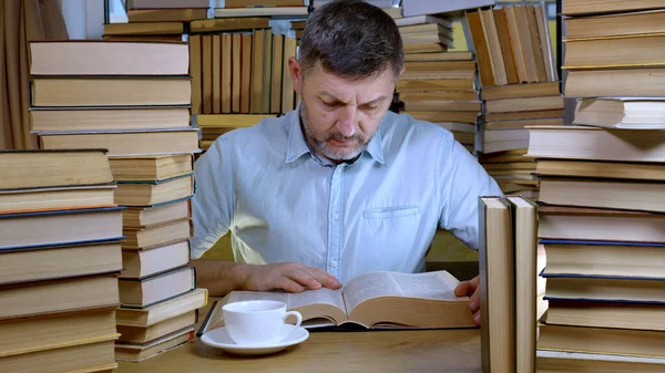 Vousatý Muž Čte Knihu Knihovně Hromádky Knih Šálek Čaje Stole — Stock fotografie