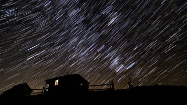Kışın dağlarda ahşap bir ev. Gece gökyüzünde yıldız izleri.