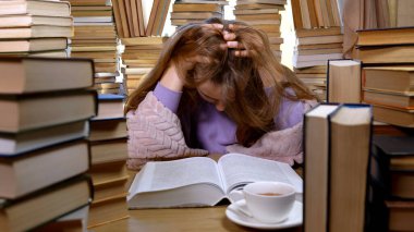 Kütüphanede bir sürü kitap arasında yorgun bir kız. Masada yığınla kitap ve çay var. Okuma ve Eğitim Konsepti.