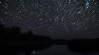 Kuyrukluyıldız şeklindeki yıldızların gece gökyüzünde gölün üzerindeki zaman çizelgeleri. Yıldızlar bir kutup yıldızının etrafında döner. Sudaki yıldızların yansıması. Efsanevi video 4K