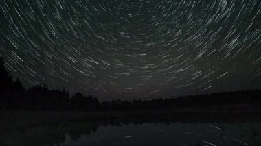 Kuyrukluyıldız şeklindeki yıldızların gölün üzerinde gece gökyüzünde süratle ilerlemesi. Yıldızlar bir kutup yıldızının etrafında döner. Sudaki yıldızların yansıması. Efsanevi video 4K