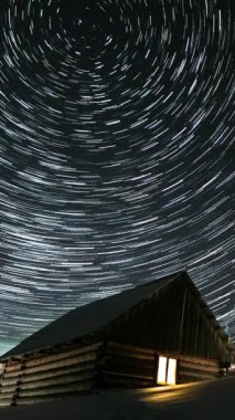 Gece gökyüzünde yıldız geçişleri, kışın dağlarda ahşap evler, kutup yıldızının etrafında dönen yıldızlar, dikey görüntüler.