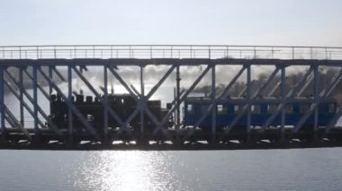 Eski bir buharlı lokomotifin nehir üzerindeki bir demiryolu köprüsünde seyahat edişinin hava görüntüsü. İnsansız hava aracı tarafından yukarıdan görülen beyaz dumanlı tarihi buhar lokomotifi.