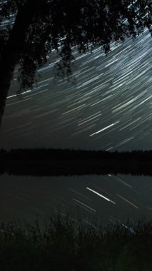 Gölün üzerindeki gece gökyüzünde yıldız izleri zaman aşımına uğradı. Sudaki yıldızların yansıması. Destansı video, dikey görüntü