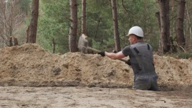 Beyaz başlıklı bir işçi aktif olarak ormanlık alanda dar bir çukur kazıyor. Çevredeki arazi, görünen ağaç kütükleri ve toprak yığınlarıyla dengesizdir..
