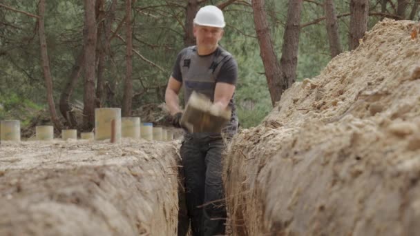 白いハード帽子をかぶった労働者は 森林地帯の狭い溝を積極的に掘っている 周囲の地形は目に見える木の茎と土のヒープで不均等です — ストック動画