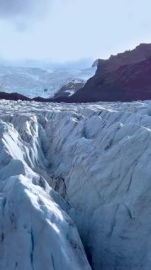 Uçsuz bucaksız bir buzulun, engebeli uçurumlarla çevrili derin mavi çatlakları. Güneş ışığı İzlanda 'daki Svinafellsjokull Buzulu' nun zıt dokularını vurgular. dikey görüntüler