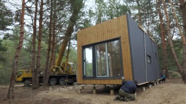 Installation of a Modern Prefabricated Cabin in a Forested Area During. The process involves precise maneuvering and placement of the structure onto a foundation amongst the tall pine trees.