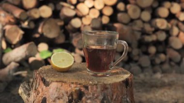 Orman ortamında bir ağaç kütüğünün üzerinde duran bir bardak çay ve bir dilim limon. Arkaplan, istiflenmiş kütüklerin bulanık bir görüntüsüdür.