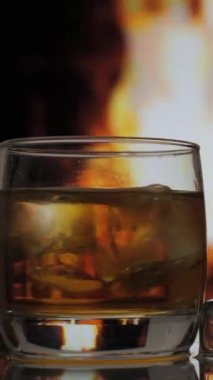 Bir bardak viski, viski, viski ya da viski şöminenin arka planında alevli bir şömine karşısında duruyor. Alkolik içki ve ev konforu. dikey görüntüler