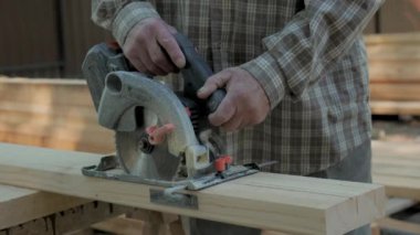 Yetenekli bir birey, sakin bir orman ortamındaki ahşap bir yapı için kereste kesmek için dairesel bir testere kullanıyor. Güneş ışığı ağaçların arasından süzülüyor, çalışma alanına yumuşak gölgeler bırakıyor. Yakın plan.