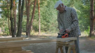 Yaşlı bir zanaatkar, sakin bir orman ortamında ahşap bir kalası ustalıkla şekillendirir. Uzun ağaçlar ve doğal ışıklarla çevrelenmiş halde, tahtayı işlemek için elektrikli bir planör kullanırken işine odaklanıyor..